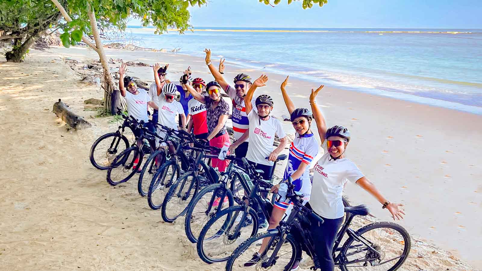 Tour group of cyclists waving to camera while on Thai beach